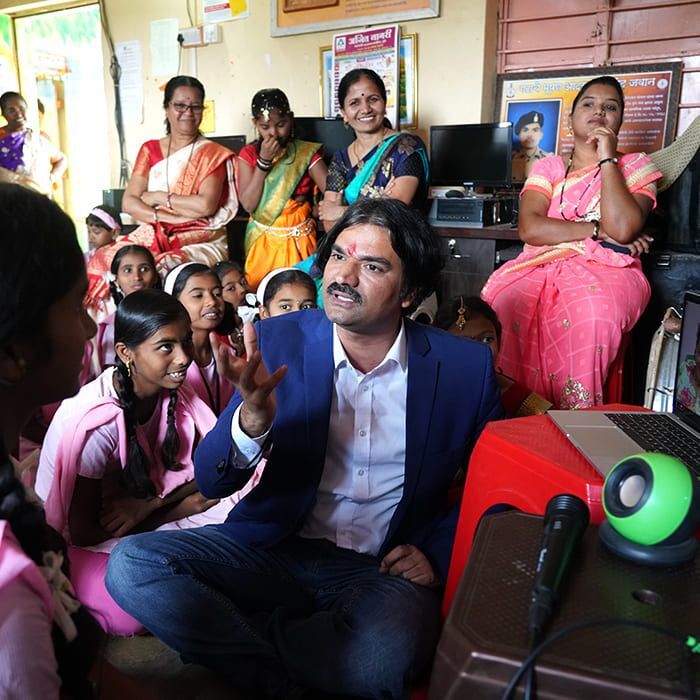 Santosh Talaghatti meeting with mothers and schoolgirls around a laptop and speaker during a community engagement session.